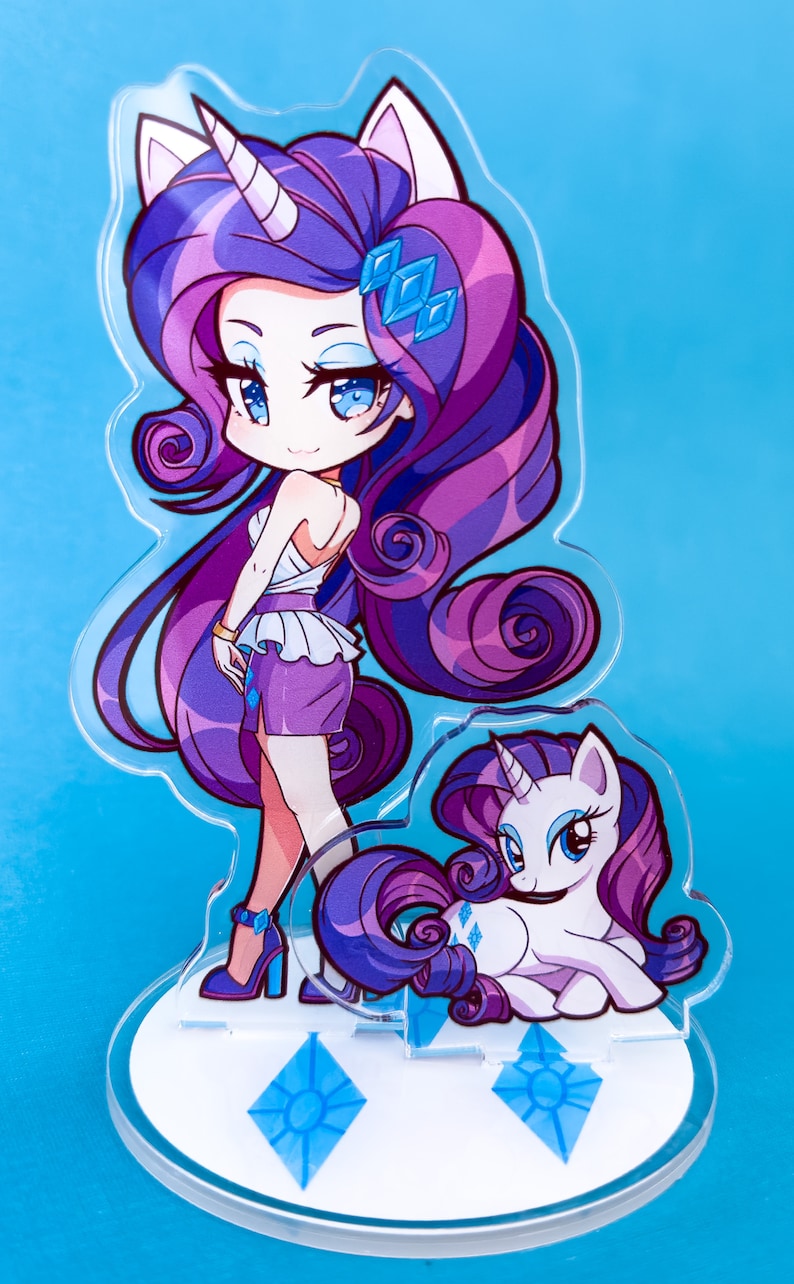 May include: A clear acrylic stand with a purple and white cartoon unicorn character. The unicorn has a blue eye and a pink horn. The stand has two blue diamond shapes on the base.