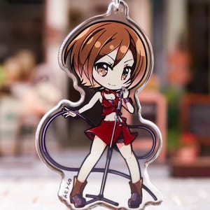 May include: A clear acrylic keychain featuring a chibi-style anime character with short brown hair, wearing a red top and skirt, holding a microphone on a stand with a black cord. The character has brown eyes and wears brown boots. Text in Japanese is visible.