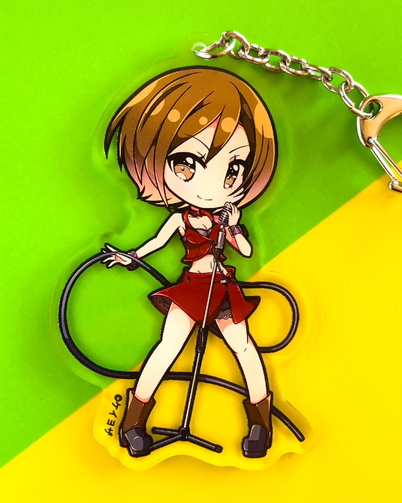 May include: An acrylic keychain featuring a chibi-style character with short brown hair, wearing a red top and skirt, holding a microphone on a stand with a black cord. The character has brown eyes and wears brown boots. The text "ケイヨザ" is printed.
