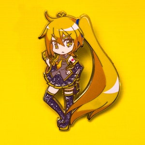 May include: A gold-colored enamel pin featuring a cartoon anime character with long yellow hair, wearing a purple and gold dress and holding a card. The character has a pink bow in her hair and blue eyes.