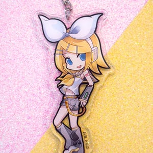 May include: A clear acrylic keychain featuring a cartoon anime character with blonde hair, a white bow, and a yellow and blue outfit. The character is holding a device with a green screen and has a red number "02" on their arm. The keychain has the text "Crypton" on the bottom.
