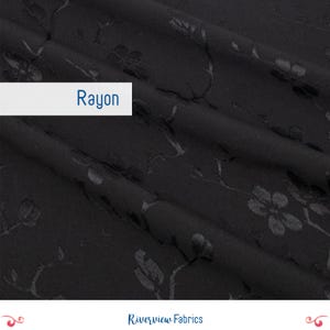 Op de afbeelding: Zwarte rayon stof met een subtiel bloemenpatroon. De stof heeft een zachte glans en een delicate textuur. Het woord "Rayon" staat op een witte rechthoek. Geschikt voor kleding en knutselprojecten.