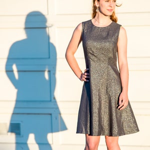 May include: A young person wearing a silver and gray sleeveless dress with a fitted bodice and a flared skirt. The person is standing in front of a white wall with a shadow behind them.