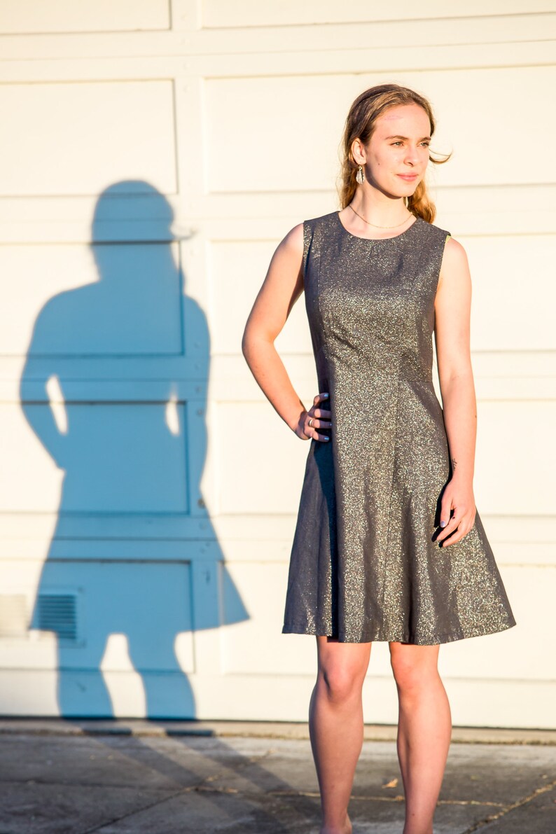 May include: A young person wearing a silver and gray sleeveless dress with a fitted bodice and a flared skirt. The person is standing in front of a white wall with a shadow behind them.