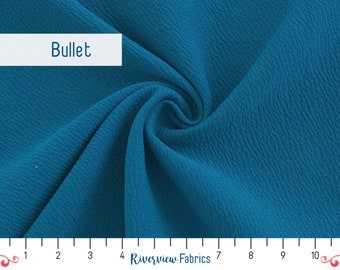 May include: A close-up of a teal blue fabric with a textured surface. The fabric is folded and draped, showing its texture and drape. A ruler is visible at the bottom of the image, with markings from 1 to 10.