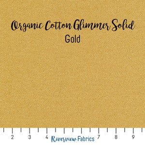 May include: A close-up of a gold organic cotton fabric with a subtle shimmer. The fabric is labeled "Organic Cotton Glimmer Solid Gold".