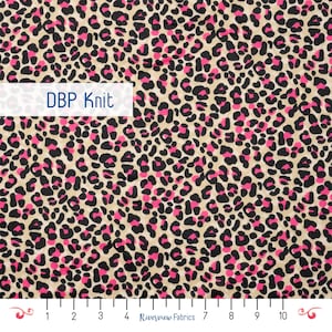 May include: A fabric with a leopard print pattern. The design features black and pink spots on a tan background. The text "DBP Knit" is displayed on a white rectangle. The fabric is marked with a ruler.