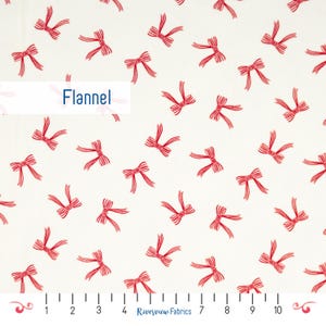 May include: White flannel fabric with a repeating pattern of red bows. The word "Flannel" is printed in blue. The fabric has a ruler along the bottom edge with Riverview Fabrics printed on it.