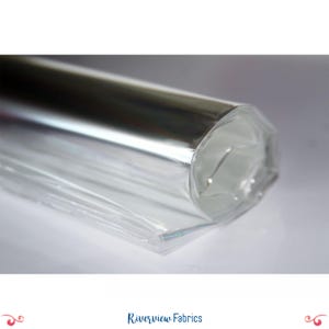 May include: A roll of shiny, silver fabric wrapped in clear plastic. The fabric reflects light, creating a bright, metallic appearance. The clear plastic wrap allows a glimpse of the fabric's texture. The Riverview Fabrics logo is at the bottom.