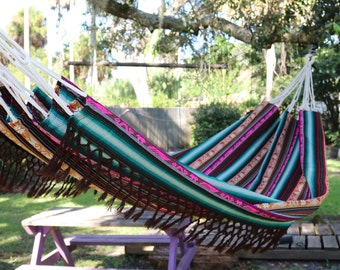 May include: A colorful striped hammock with brown fringe hangs between two trees. The hammock is made of woven fabric and has a bright pink stripe running through the center.