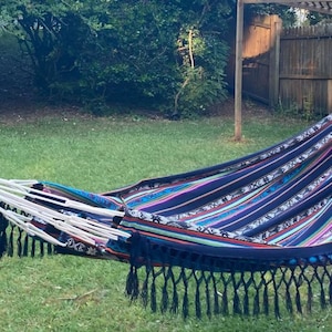 May include: A colourful hammock with black fringe hangs between two trees in a grassy backyard. The hammock is made of woven fabric with stripes of blue, pink, green, and white.