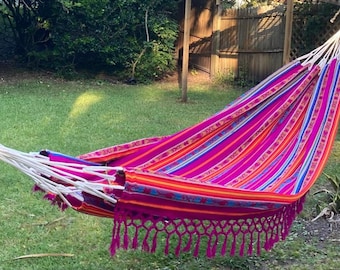 May include: A pink, orange, and blue striped hammock with fringe. The hammock is hanging between two trees in a grassy backyard.