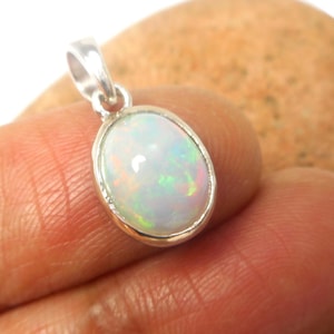 May include: Oval-shaped opal pendant with a silver-colored setting. The opal displays a play of color with iridescent flashes of green, blue, and yellow. The pendant is suspended from a small loop.