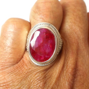 May include: A silver ring with a large oval-shaped ruby gemstone. The ring is set in a silver band with a decorative border.