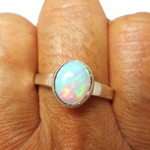 Può includere: Anello in argento con un'opale ovale. L'opale mostra giochi di colore con lampi iridescenti blu, verdi e rossi. L'anello è su un dito su sfondo bianco.