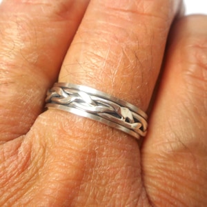 May include: A silver ring with a braided design. The ring is worn on the finger of a person's hand.
