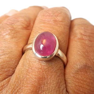 May include: A silver ring featuring a smooth, oval-shaped, translucent pink gemstone set in a simple bezel. The stone has a polished, rounded cabochon surface that reflects light, and the band is a plain, polished silver metal.