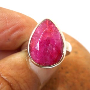 May include: A teardrop-shaped faceted ruby gemstone set in a polished silver ring band. The stone features a deep magenta-pink hue with a textured, multi-faceted surface that reflects light, held within a simple, smooth silver metal bezel setting.
