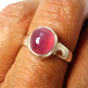 May include: A silver ring with a large, oval, cabochon-cut, pink gemstone. The ring's band is smooth and polished, and the gemstone is set in a bezel setting. The ring is shown on a finger.