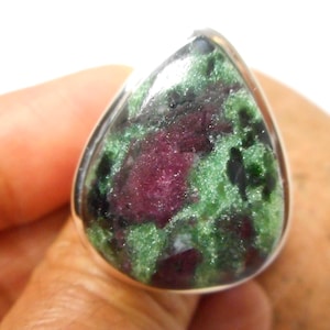 Puede incluir: Un anillo en forma de lágrima con una piedra preciosa natural de rubí zoisita. La piedra muestra una mezcla de colores rojo oscuro, verde y negro, engastada en una banda plateada. El anillo es sostenido por una mano.