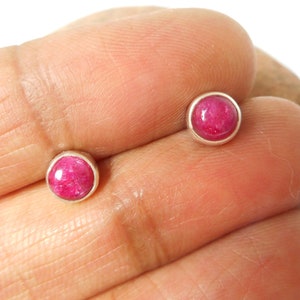 May include: A pair of round stud earrings featuring a vibrant pink gemstone set in a silver bezel. These small, delicate earrings are suitable for daily wear. The gemstones have a slightly textured appearance.