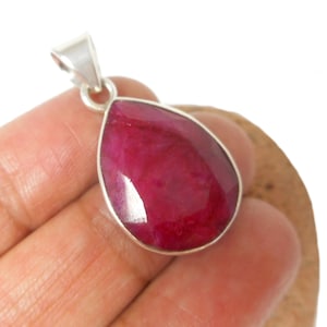 May include: A teardrop-shaped pendant featuring a faceted, deep red gemstone. The gem is set in a silver-coloured metal frame and has a loop at the top for a chain. The pendant is held in a hand against a neutral background.
