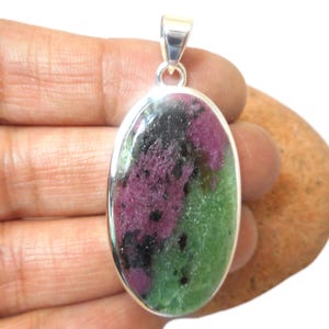 May include: Oval pendant with a silver-coloured bail. The pendant features a natural stone with a mix of colours: deep red, green, and black. The stone is set in a polished silver frame. The pendant is held in a hand.