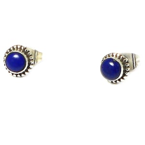 May include: A pair of stud earrings with a round, deep blue gemstone set in silver-coloured metal. The metal setting has a beaded border. The earrings are shown against a white background.