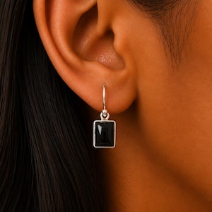 May include: A silver earring with a square, black gemstone. The earring hangs from a small hoop. The earring is shown on a person's ear, with dark hair visible.