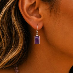 May include: A silver drop earring featuring a rectangular purple amethyst gemstone set in a polished metal frame. The earring hangs from a simple silver hook, showcasing a smooth, deep violet stone that catches the light with a subtle shine.
