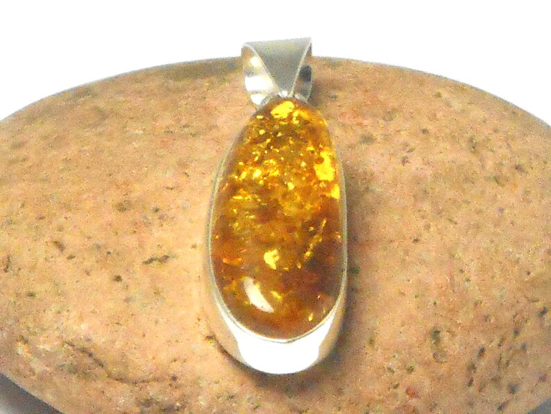 May include: A teardrop-shaped amber pendant set in a silver bezel. The amber is a rich, golden color with flecks of brown and yellow.