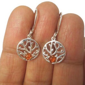 May include: A pair of silver-colored earrings. Each earring features a circular design with a tree-like pattern and a small, orange-colored stone in the center. The earrings are held up by a silver hook.