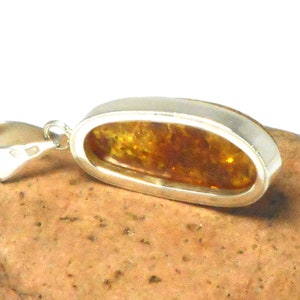 May include: A silver pendant with a large oval amber stone. The amber is a warm honey color with inclusions. The pendant is on a brown rock.