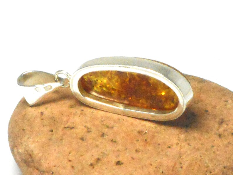 May include: A silver pendant with a large oval amber stone. The amber is a warm honey color with inclusions. The pendant is on a brown rock.