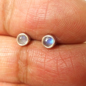May include: A pair of small, round stud earrings. Each earring has a small, iridescent, blue and white gemstone set in a silver bezel. The earrings are shown against a skin-toned background.