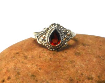 Può includere: Un anello d'argento con una pietra rossa a forma di pera incastonata in una montatura d'argento dettagliata.