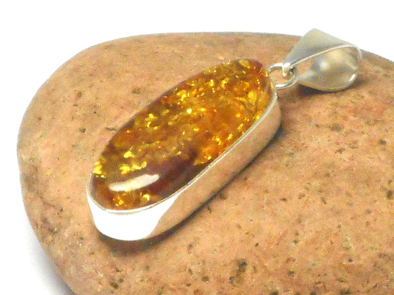 May include: A silver pendant with a large oval amber stone. The amber is a deep yellow color with flecks of gold throughout. The pendant is on a brown rock.