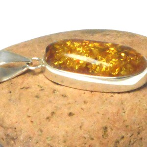 May include: A silver pendant with a large oval amber stone. The amber is a deep golden color with flecks of brown and black. The pendant is on a brown rock.
