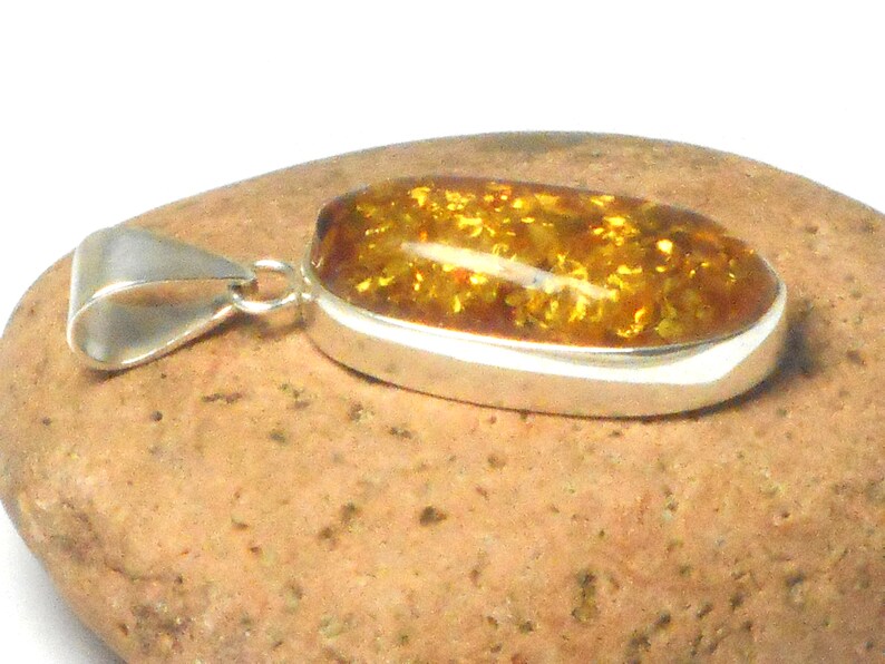 May include: A silver pendant with a large oval amber stone. The amber is a deep golden color with flecks of brown and black. The pendant is on a brown rock.
