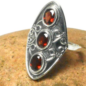 May include: A silver-colored ring with three oval, deep red gemstones. The ring has an elongated, shield-like face with ornate scrollwork and beaded edges. The gemstones are set in individual bezels.