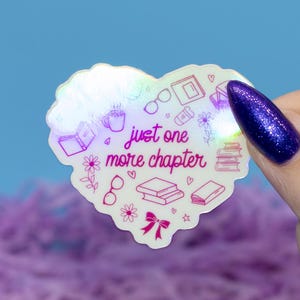 May include: A heart-shaped sticker with the text "just one more chapter". The sticker has illustrations of books, glasses, and flowers. The sticker has a holographic, iridescent finish. The background is blue and purple.