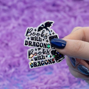 May include: Two iridescent holographic stickers with the text "Books with Dragons" and a black silhouette of a dragon. The stickers are held in a hand with dark blue nail polish.
