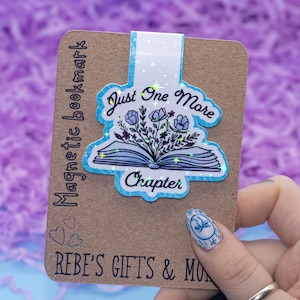 Puede incluir: Un marcapáginas magnético azul y blanco con un diseño de libro y flores. El marcapáginas tiene el texto "Just One More Chapter".
