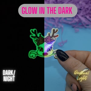 May include: A glow-in-the-dark reindeer sticker with a pink bow. The sticker is shown in both daylight and dark conditions. "Glow in the Dark" and "Daytime/Light" are written on the image.