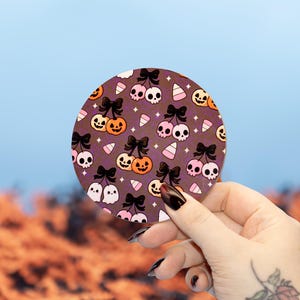 Può includere: Un sottobicchiere rotondo con un motivo a tema Halloween. Il design presenta teschi, zucche, fantasmi e caramelle a forma di pannocchia, tutti con fiocchi neri, su uno sfondo viola scuro. Il sottobicchiere è tenuto contro uno sfondo sfocato arancione e blu.