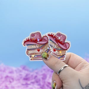 May include: Two stickers depicting a pink cowboy hat on a stack of books. The hat has a red brim and star decorations. The books have a heart pendant and star accents. The background is a light blue sky.