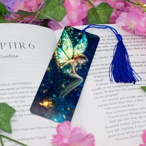 Puede incluir: Un marcapáginas de metal azul y verde con un hada con las alas extendidas. El hada está sentada en una rama con un fondo estrellado. El marcapáginas tiene un borla azul.