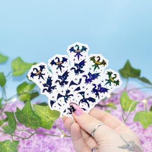 May include: A selection of dragon stickers with a holographic glitter finish. The stickers depict different dragon designs in shades of blue, green, and purple. The stickers are held against a backdrop of purple paper and green leaves.