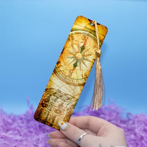 Może przedstawiać: Złota zakładka z vintage'owym wzorem kompasu i skrzyni skarbów. Zakładka ma złoty frędzl.