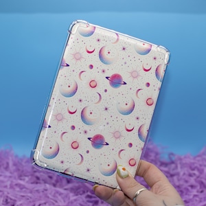 Può includere: Una custodia trasparente per tablet con un motivo celeste di pianeti, lune e stelle nei toni del rosa, viola e blu. La custodia ha un paraurti protettivo. Lo sfondo è azzurro.